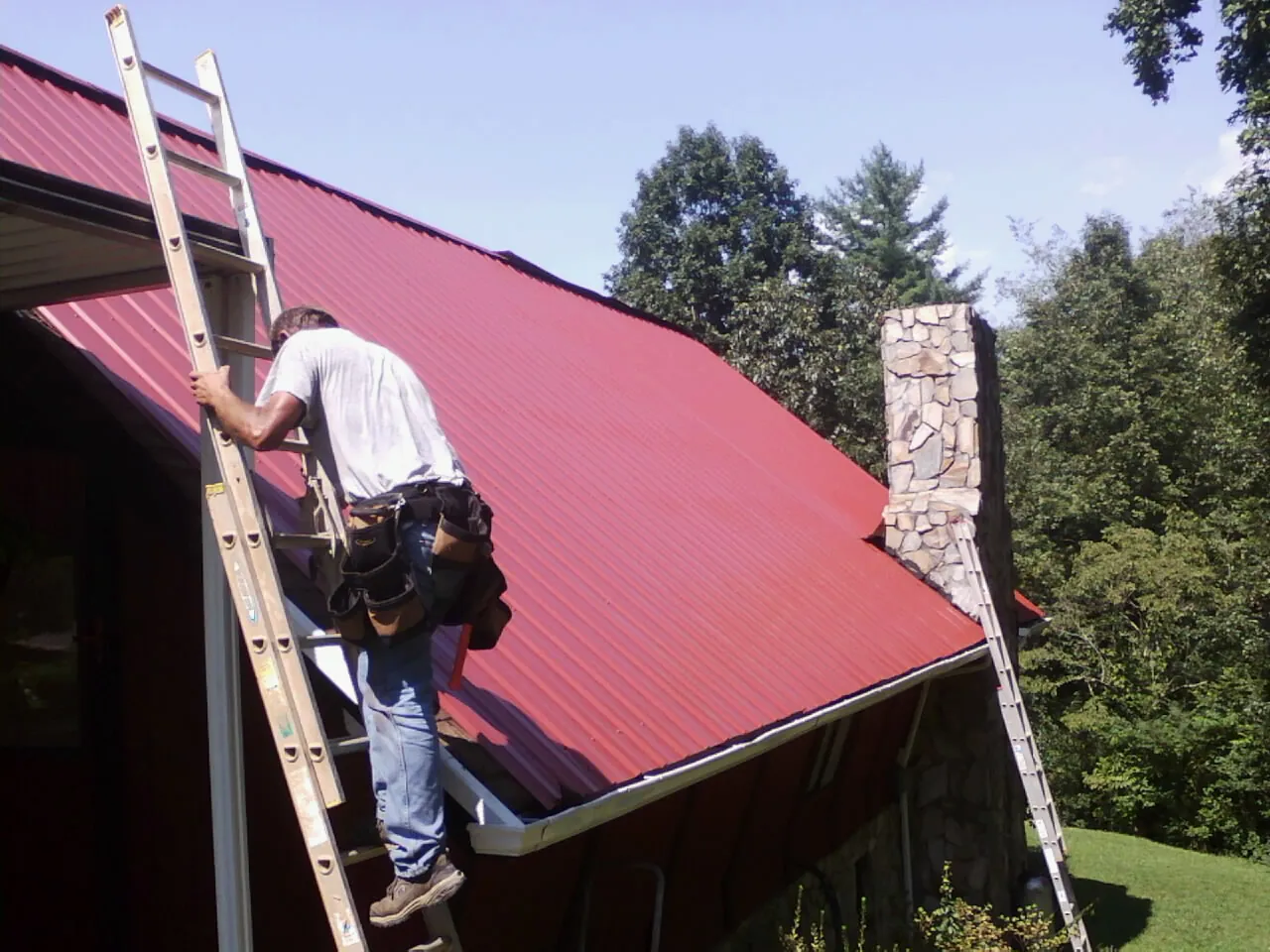 Roofer on a ladder beside a red metal roof and stone chimney — Soffit & Fascia Repair by Roofing Brothers in Wapakoneta